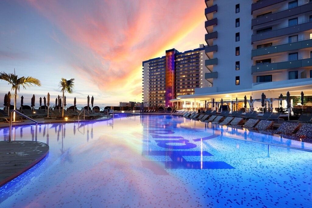 Swimming pool illuminated at sunset in a hotel in Tenerife, surrounded by sun loungers and palm trees.
