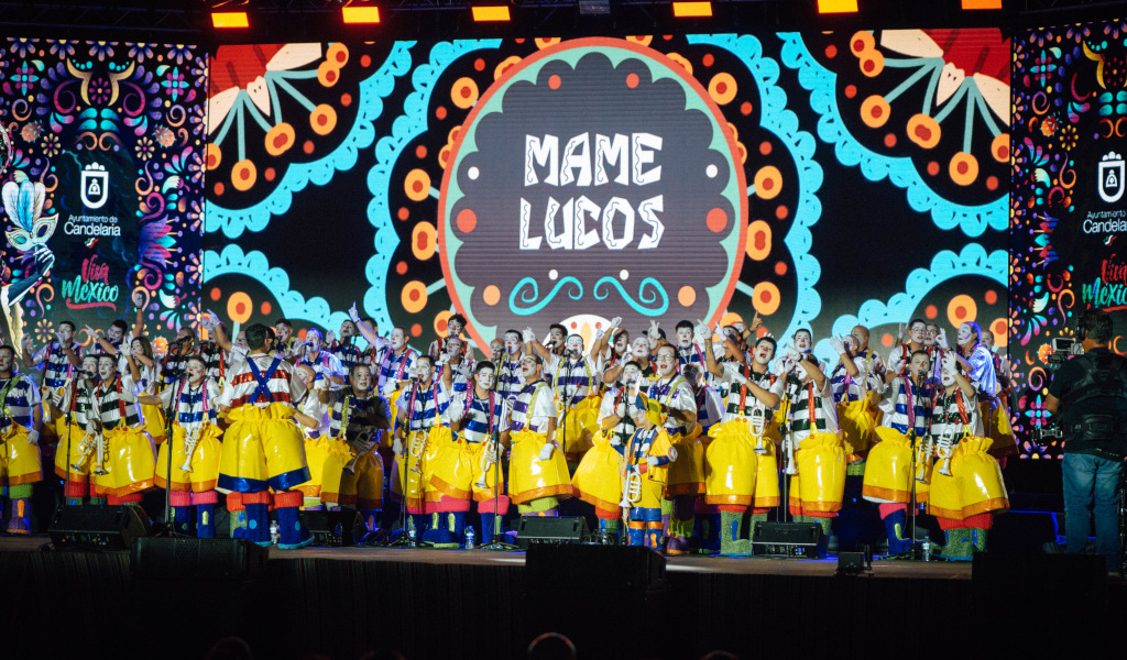 Group of people on the stage of Candelaria, with colorful background and text Mame Luicos.