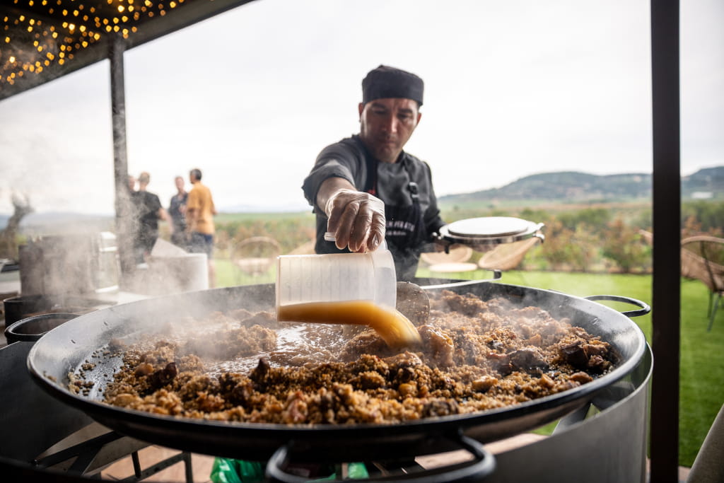 Cook pouring broth into a large outdoor paella in Tenerife.