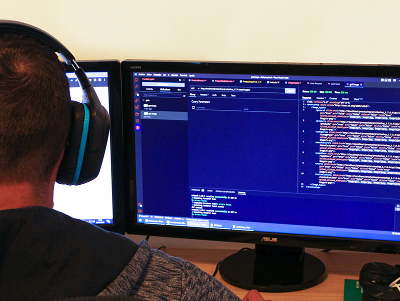 Man in headphones coding on dual monitors, displaying JSON and query parameters.