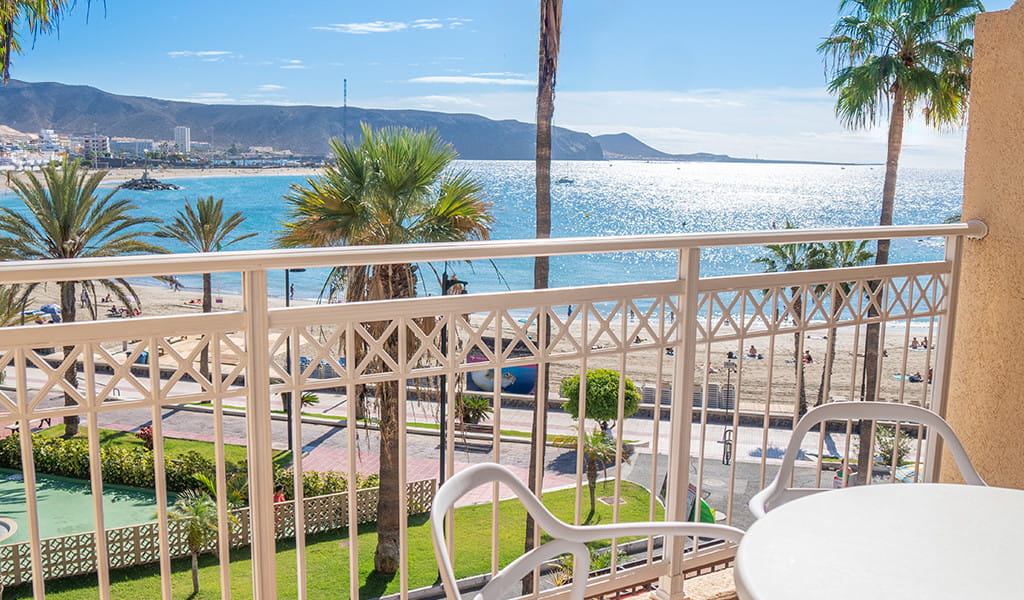 View from a balcony overlooking the beach in Tenerife, with palm trees and ocean.