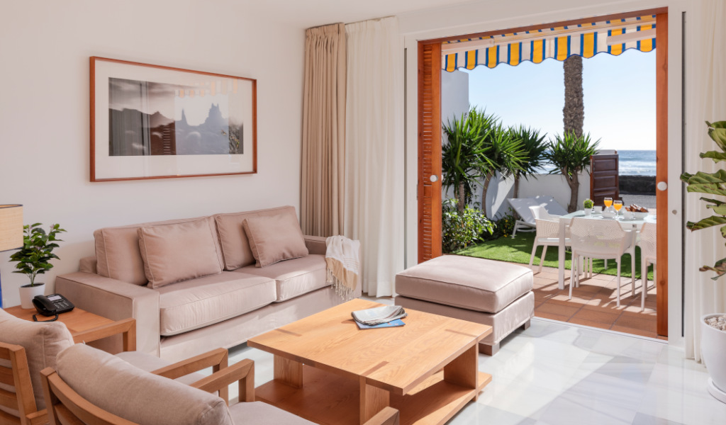 Living room with beige sofa and coffee table, view of Tenerife garden and sea through open doors.
