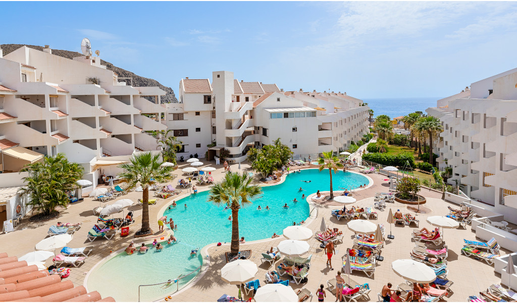 People enjoy a pool at a resort in Los Cristianos, Tenerife, with sea view in background.