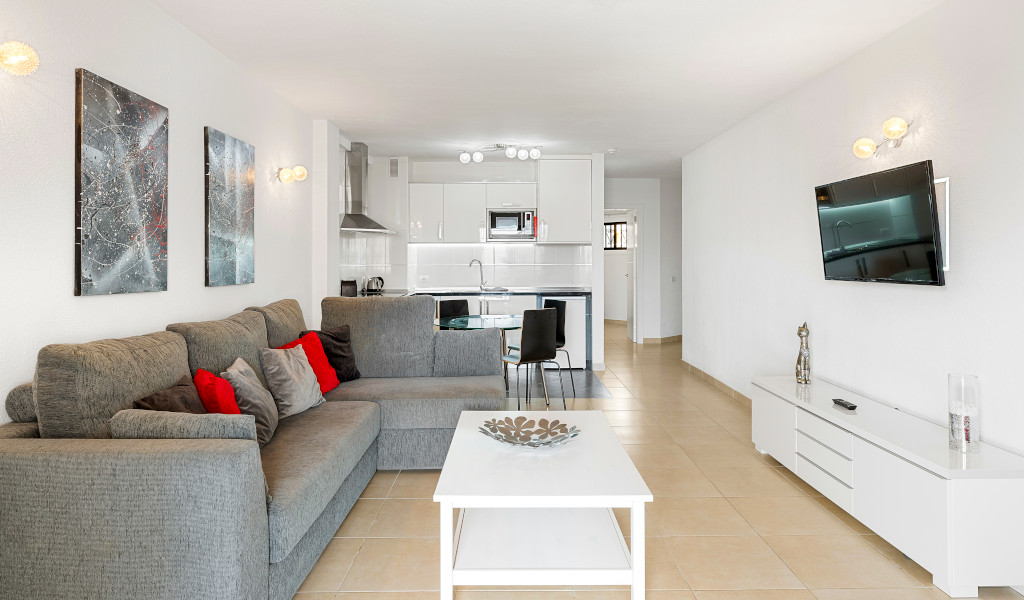 Living room with grey sofa, TV, and kitchen in a Tenerife apartment.