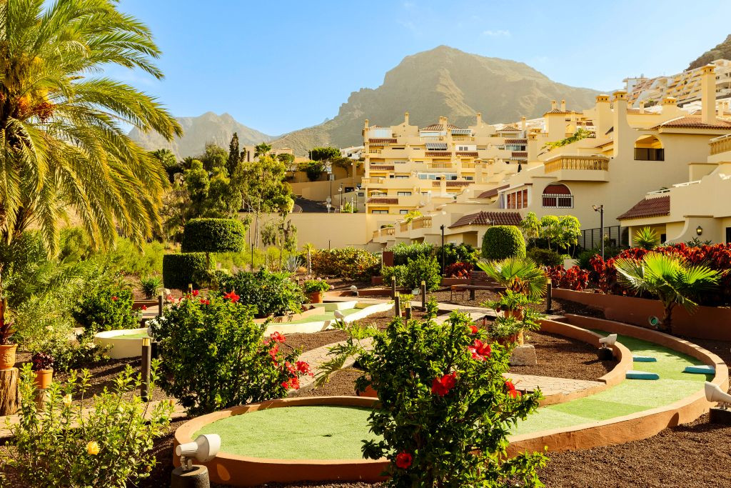 Mini-golf course surrounded by green shrubs and palm tree, with Costa Adeje apartments in background.