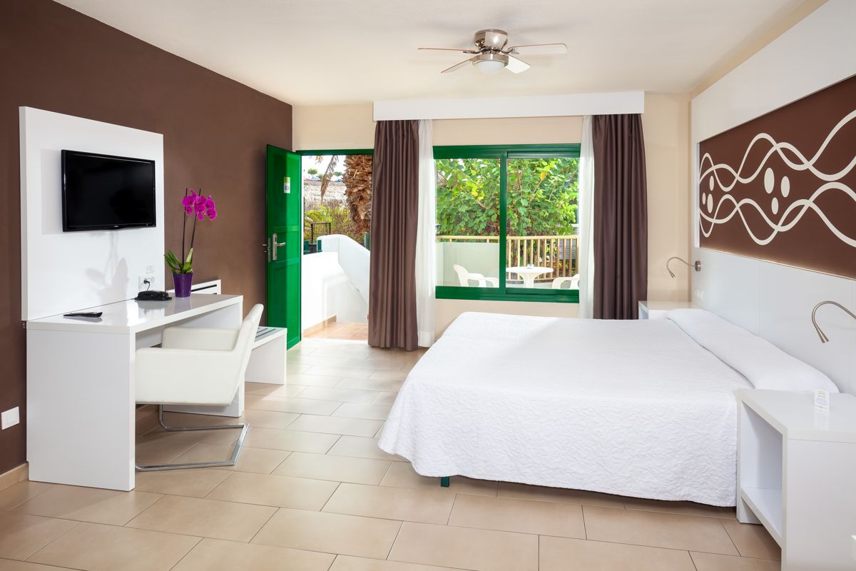 Hotel room in Tenerife with bed, desk, TV, green door, and window facing garden.