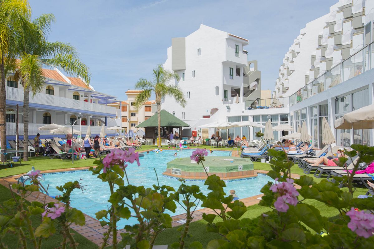 Outdoor pool area with people lounging and swimming, hotel in Tenerife background.