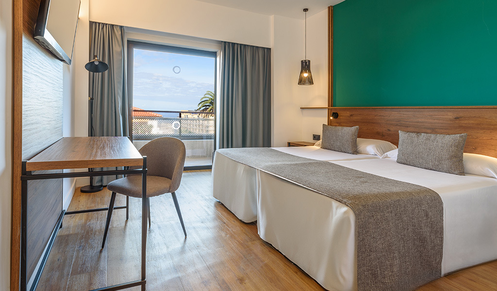 Hotel room in Tenerife with two beds, desk, chair, and view of ocean and palm trees.