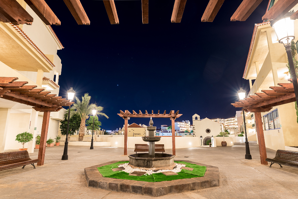 Courtyard with fountain at night in Tenerife, surrounded by benches and palm trees.