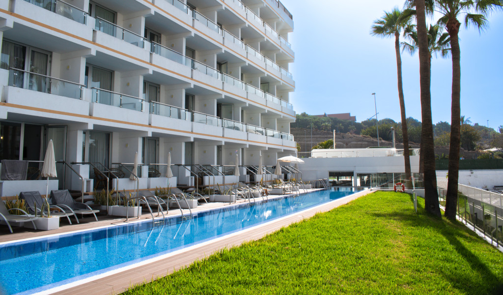 Hotel with pool and sun loungers in Tenerife, surrounded by grass and palm trees.