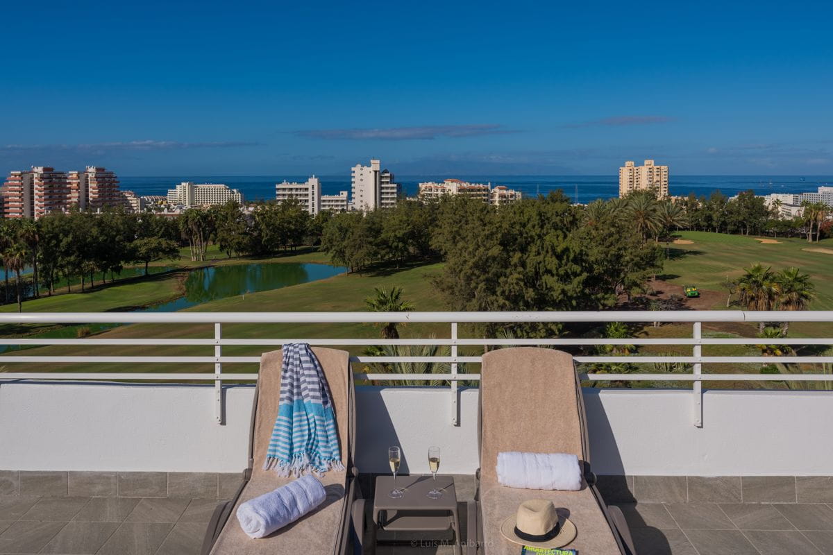 Two lounge chairs overlook a golf course and ocean view in Tenerife, with buildings in the background.