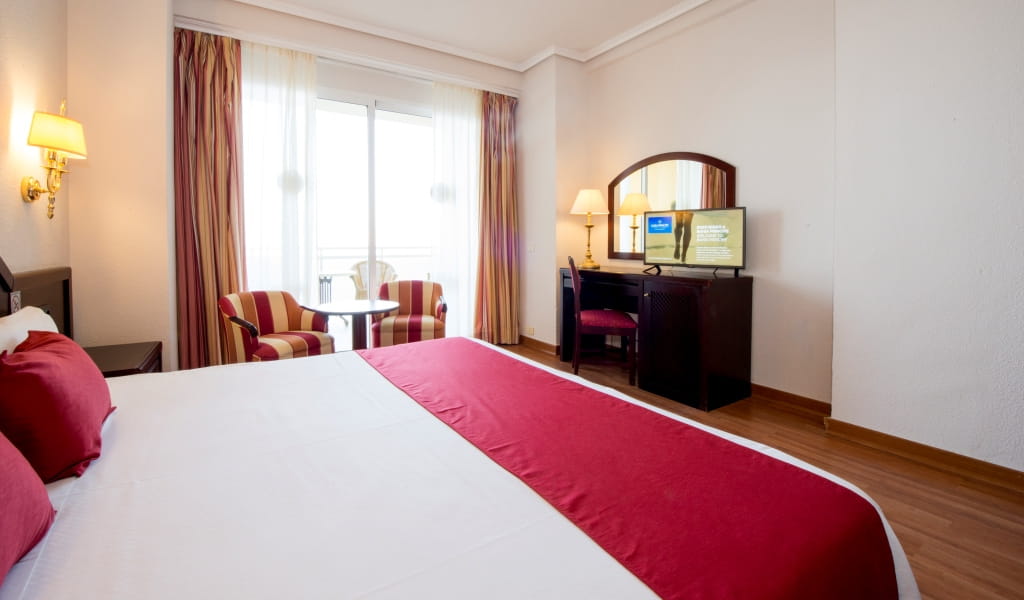 Hotel room with red bedding, desk with TV, and balcony in Tenerife.