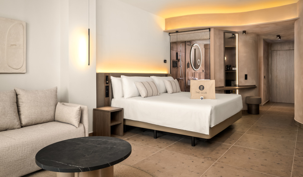 Modern hotel room with bed, sofa, table, and The Club branded bag near mirror in Tenerife.