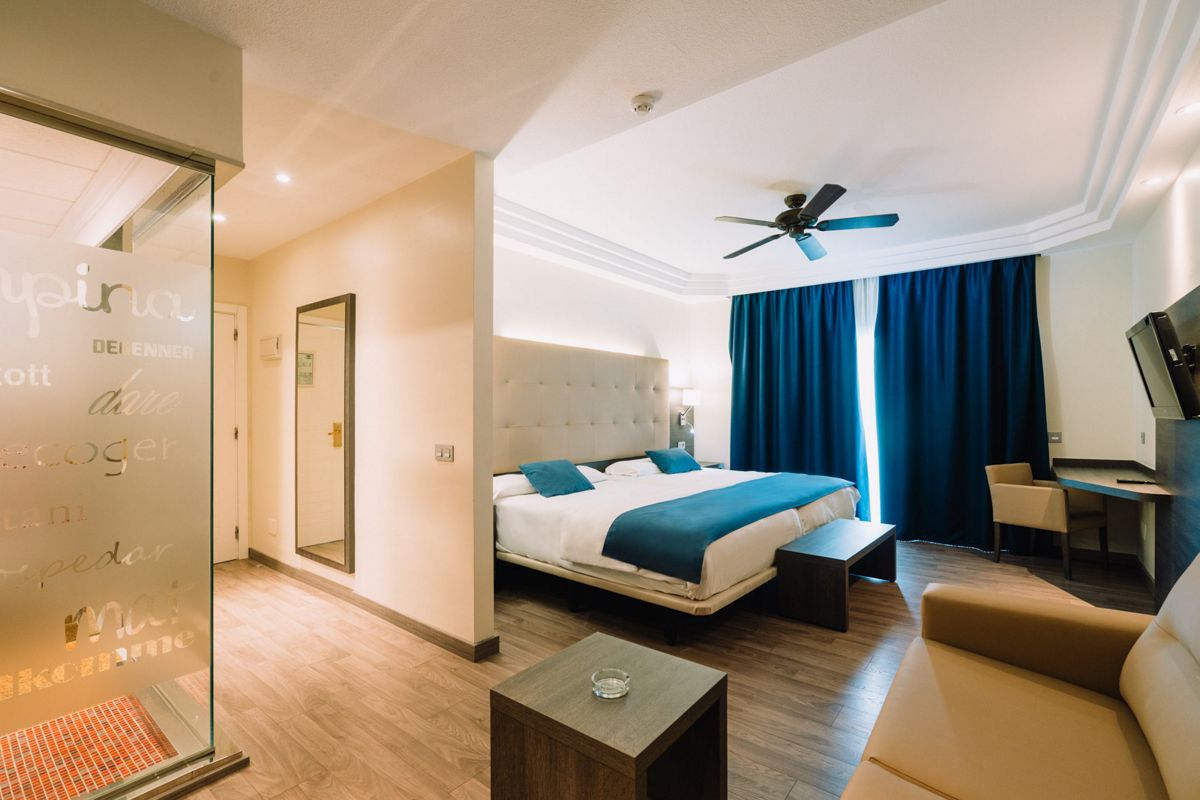 Hotel room in Tenerife with double bed, blue curtains, and seating area. Room door and mirror visible.