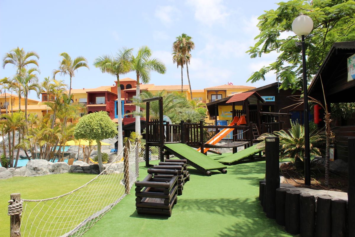 Children's playground with slides and palm trees, Tenerife resort in the background.
