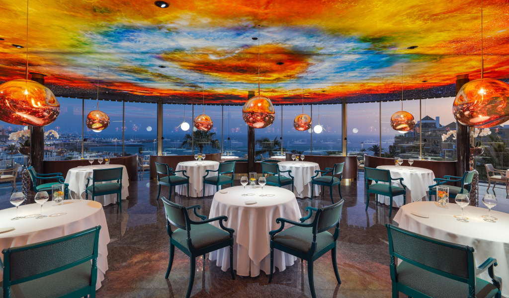 Restaurant with colorful ceiling and sea view in Tenerife, round tables set for dining.