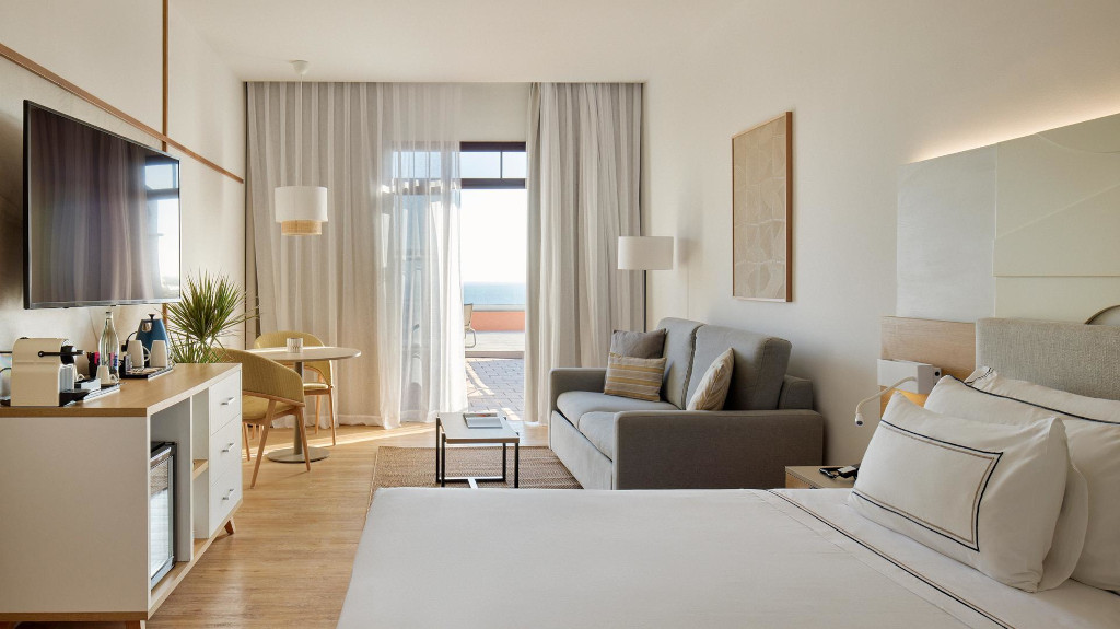Spacious hotel room in Tenerife with ocean view, TV, bed, sofa, and desk.