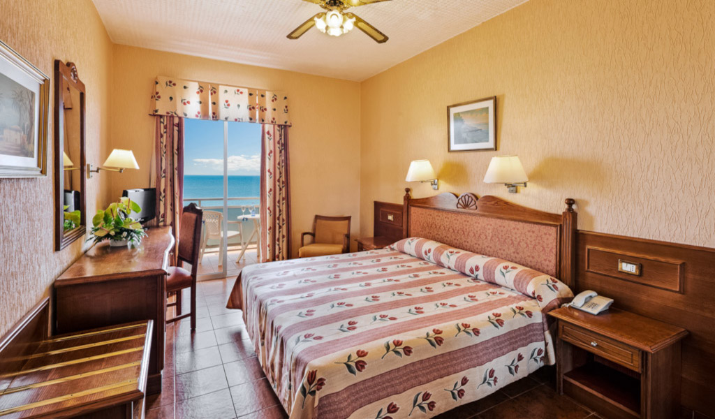 Bedroom with floral bedspread, desk, and sea view from window in Tenerife hotel.