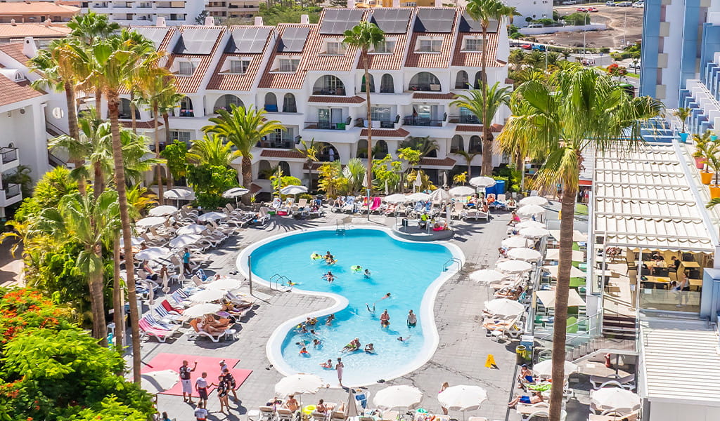 Outdoor pool with people swimming at a Tenerife resort, surrounded by palm trees and sun loungers.