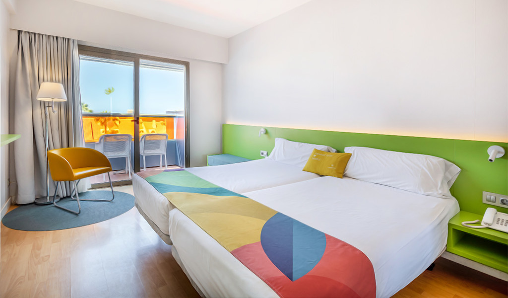 Twin beds with colorful bedding in a room with balcony and chair in Tenerife hotel.