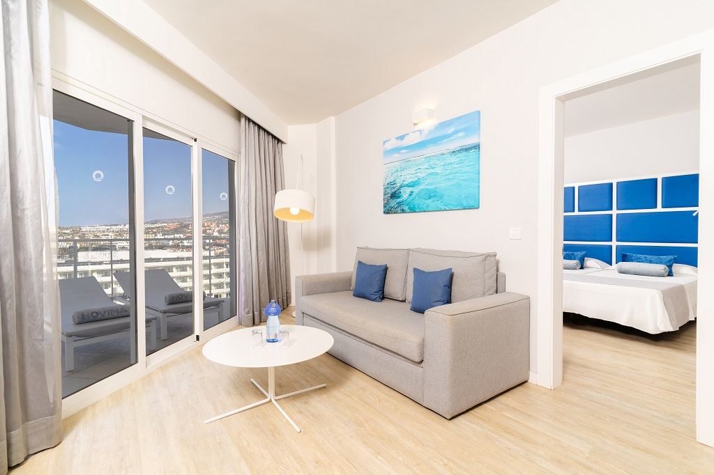 Living room with sofa, balcony view of Costa Adeje, and adjoining blue-themed bedroom.