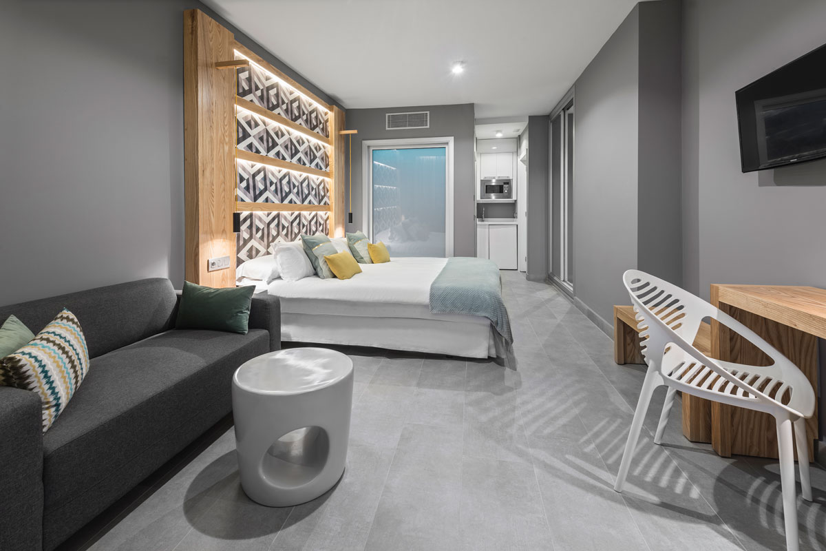 Modern hotel room in Tenerife with double bed, sofa, desk, and kitchenette.