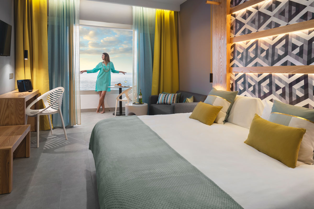Hotel room with ocean view in Tenerife, woman by window, bed in foreground.