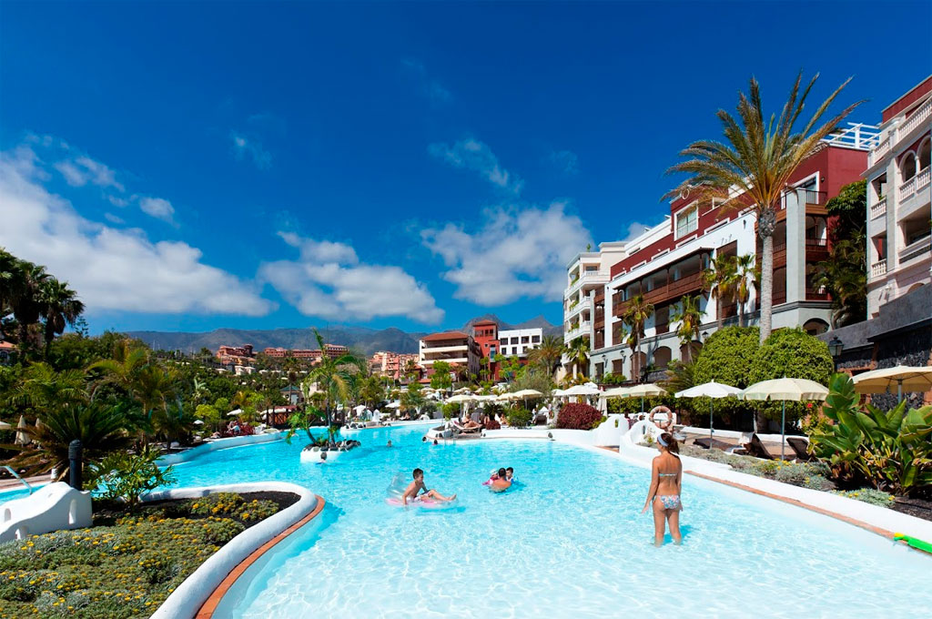 Outdoor swimming pool with people, surrounded by palm trees and a hotel in Tenerife.