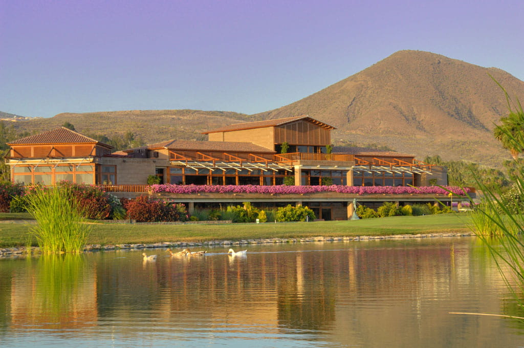 Modern clubhouse surrounded by greenery and mountains in Tenerife, reflected in a pond.