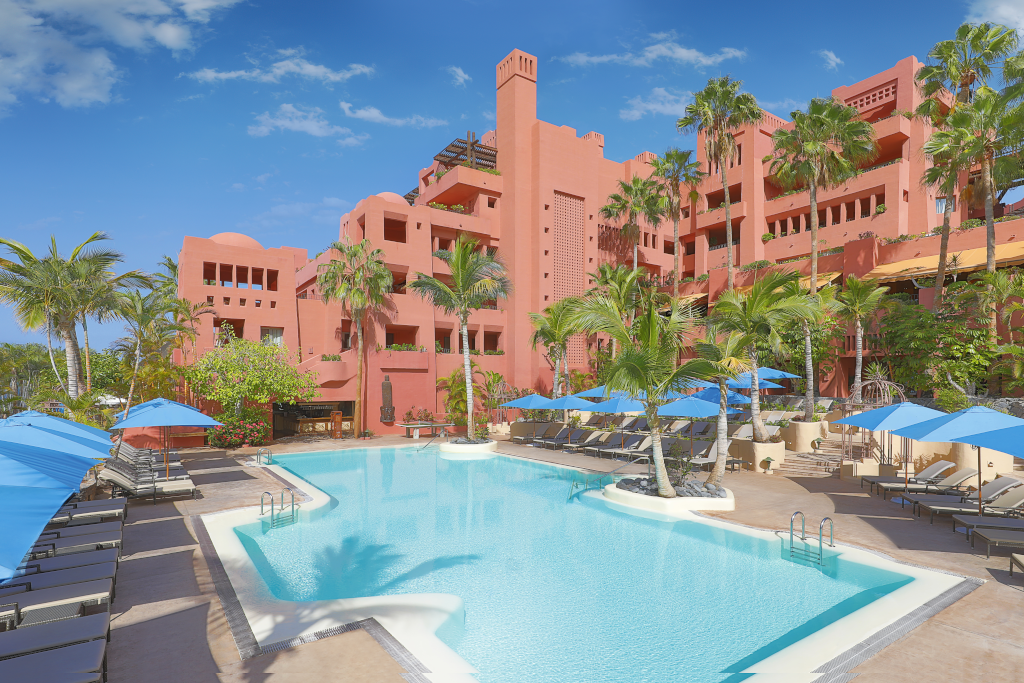 Outdoor pool area with blue umbrellas and palm trees at a pink hotel in Tenerife.