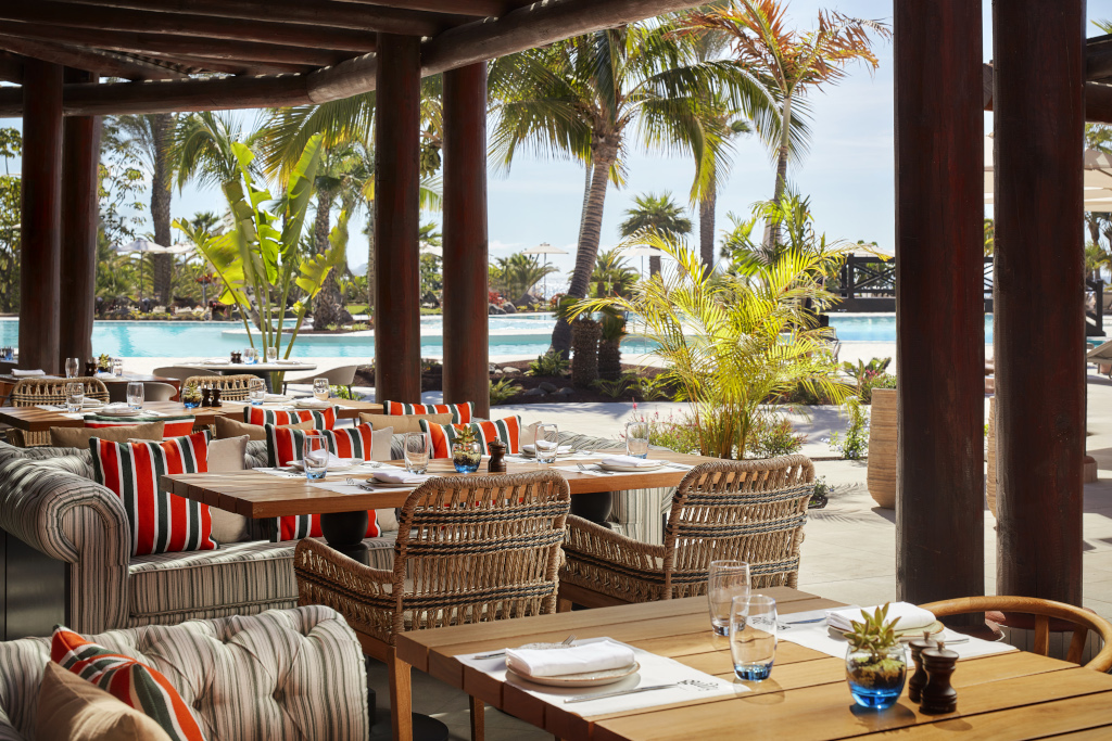 Outdoor restaurant seating with pool and palm trees at a Tenerife resort.