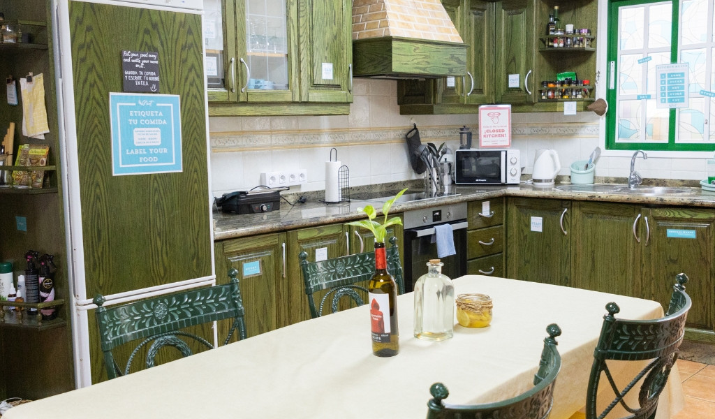 Kitchen in Tenerife hostel with green cabinets, table, bottles, and visible signs.