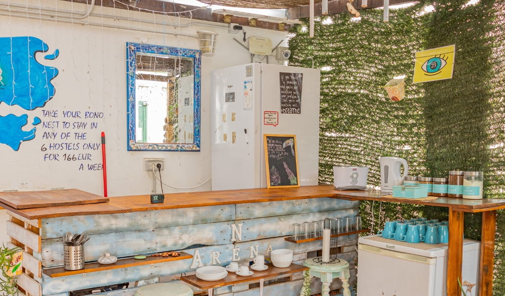 Kitchen counter with blue cups, a fridge, and a wall sign offering hostel stays for 166 EUR a week in Tenerife.