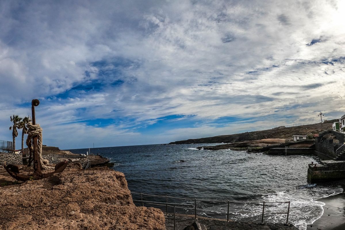 Rusty anchor on rocky coast overlooking ocean in Tenerife, with cloudy sky.