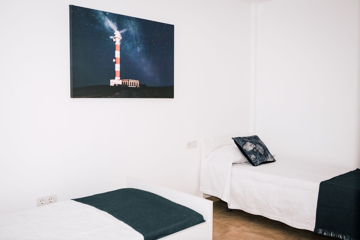 Two beds with dark blankets, painting of a lighthouse on La Palma wall above.