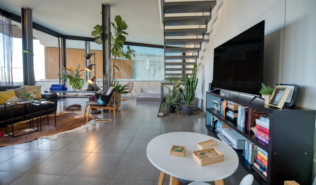 Modern living room in Tenerife with large windows, plants, black couch, and a staircase.