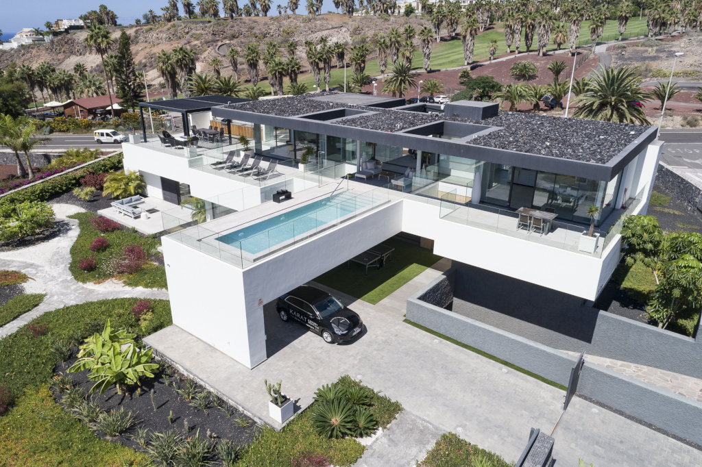 Modern villa with pool and garden in Tenerife, surrounded by lush greenery and a parked car.