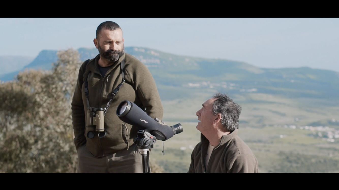 Two men with binoculars and telescope in mountainous landscape.