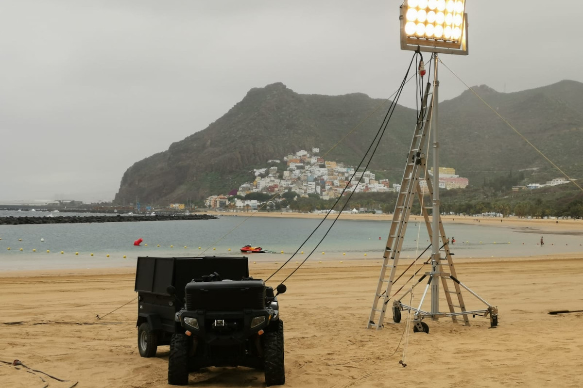Lights and vehicle on the beach of Las Teresitas, Tenerife, with houses and mountains in the background.