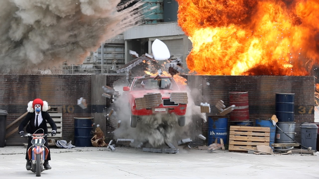 Motorcyclist with clown helmet and red car in rear explosion.