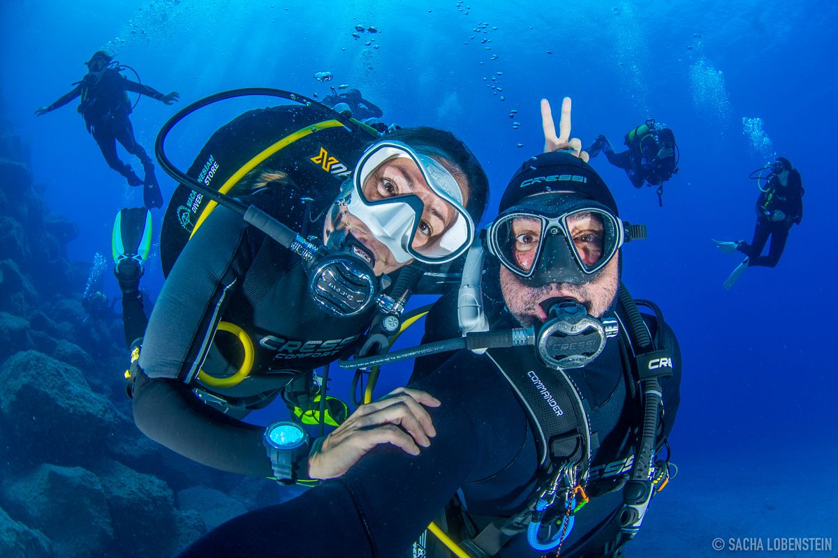 Divers smiling underwater in Tenerife with others in the background.