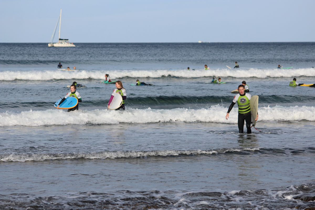 Surfers with boards in Playa de Las Américas, Tenerife, with a sailboat in the background.