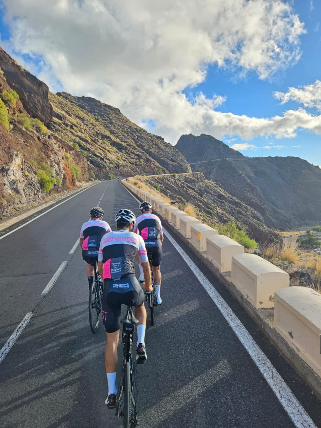 Cyclists climbing a mountainous road in Tenerife under partly cloudy skies.