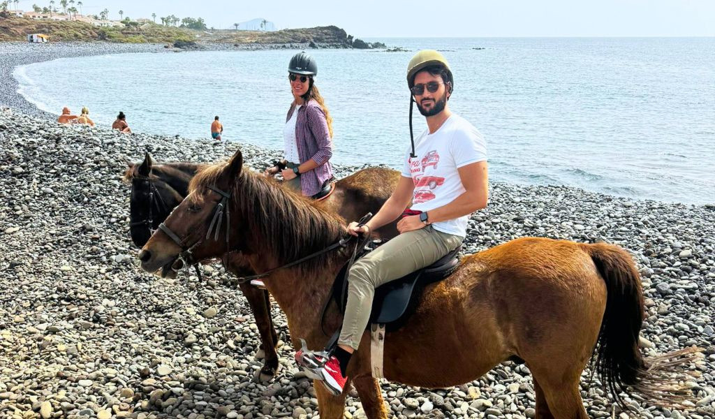 People riding horses on the pebble beach of Tenerife, with the sea in the background.