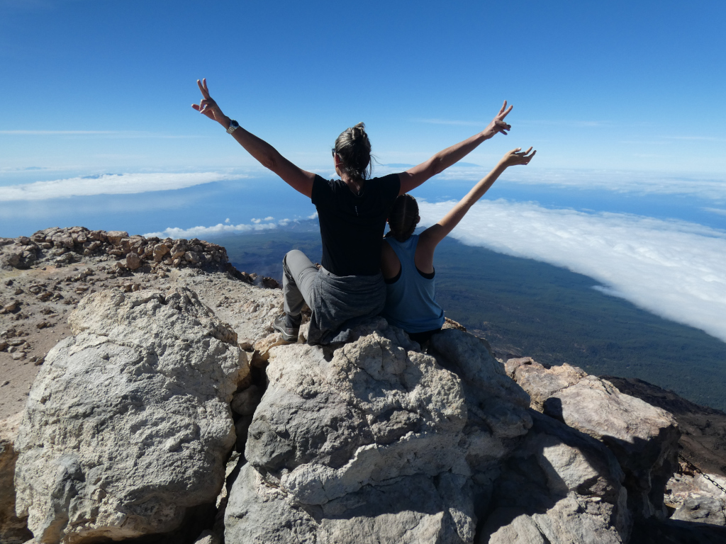 People raising their arms at the top of Mount Teide in Tenerife, overlooking the cloudy landscape.
