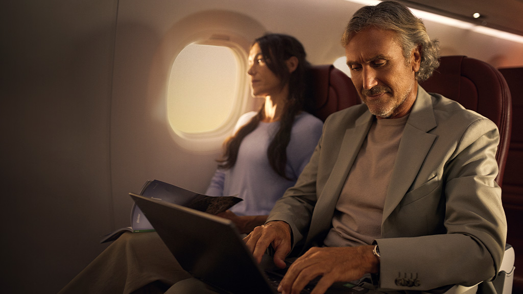 Person using laptop on a plane, accompanied by passenger reading.