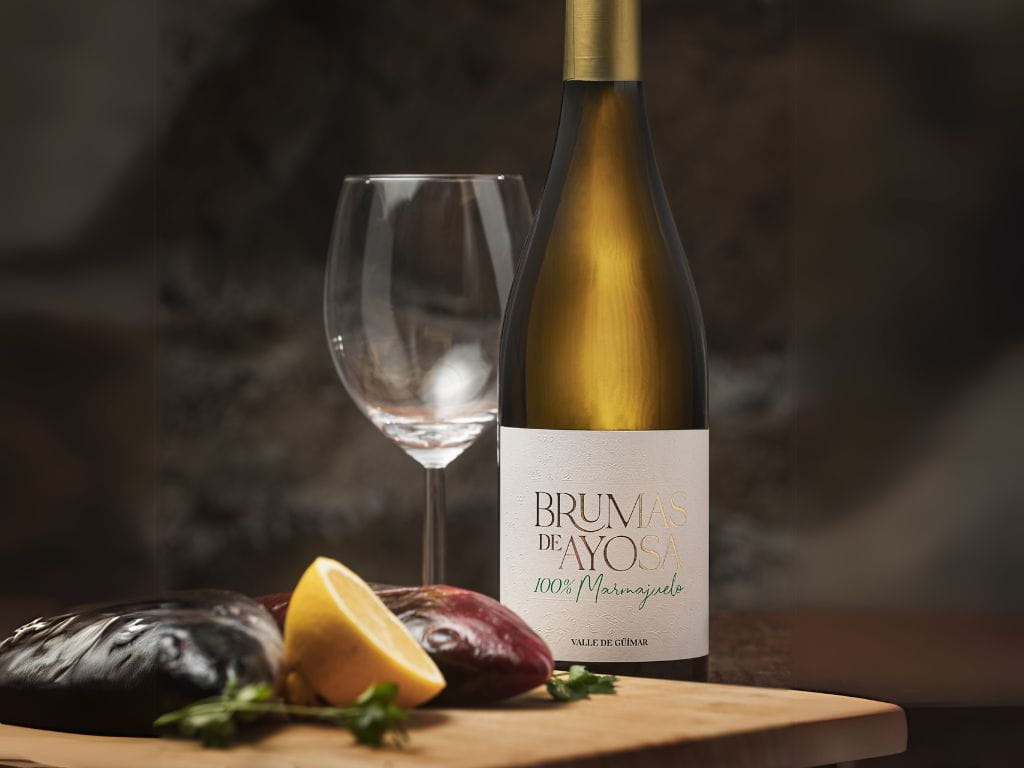 Bottle of Brumas de Ayosa wine with empty glass, lemon and aubergines. Text: 100% Marmajuelo. Güímar Valley.