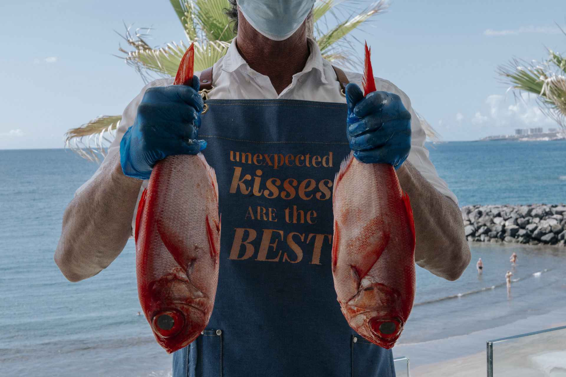 Person holding two fish with apron saying unexpected kisses are the best in Tenerife.