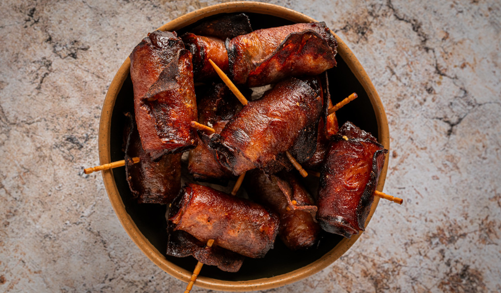 Bacon-wrapped sausages with chopsticks on stone background.