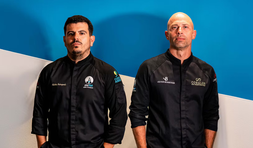 Two chefs in black uniforms pose in front of a blue and white background in Tenerife.
